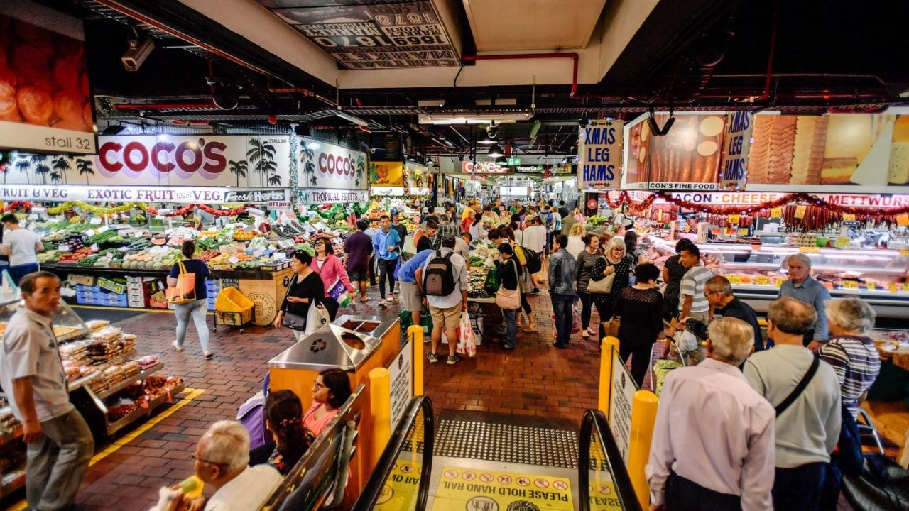 Shoppers walking through a bustling Adelaide Central Market filled with local produce and various stalls.